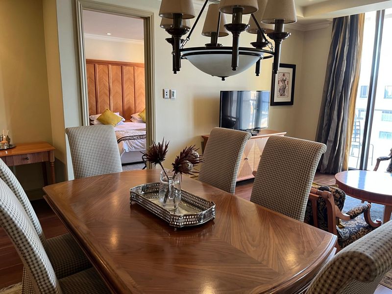 A well-maintained dining area featuring hardwood flooring, elegant cove ceiling detailing, and a central chandelier, with an adjacent bedroom visible through the doorway.