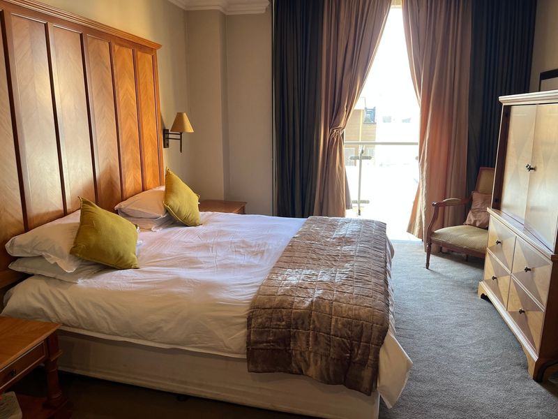 A well-lit bedroom featuring a large built-in wooden headboard, carpeted flooring, and direct access to a private balcony through sliding glass doors.