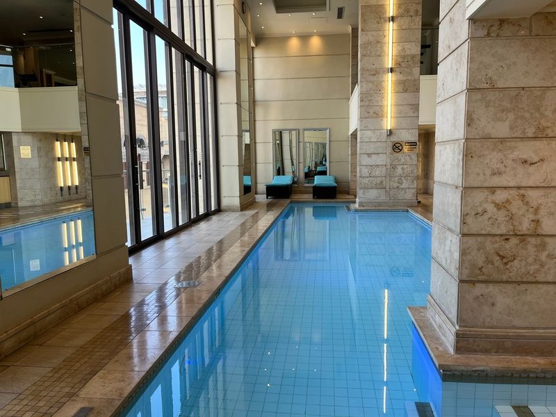 A luxurious indoor swimming pool area featuring stone-clad columns, high ceilings with recessed lighting, and large glass doors providing natural light.