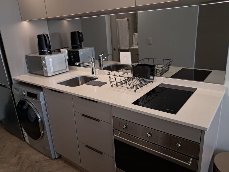 Modern compact kitchen featuring sleek cabinetry, stone countertops, integrated oven, induction cooktop, and a mirrored splashback with under-cabinet lighting.