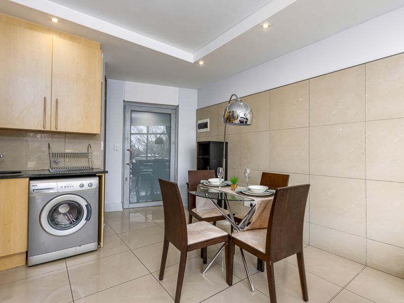 Open-plan kitchen and dining area featuring light wood cabinetry, integrated laundry appliance, tiled flooring, and a modern tiled feature wall.