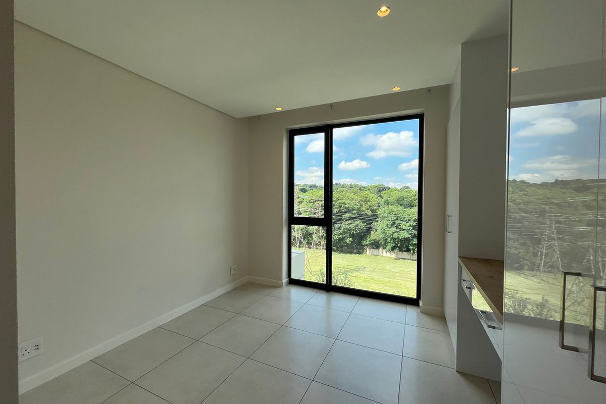 A bright, modern, and empty bedroom featuring neutral tiled flooring, built-in cupboards, and a large window offering scenic green views.