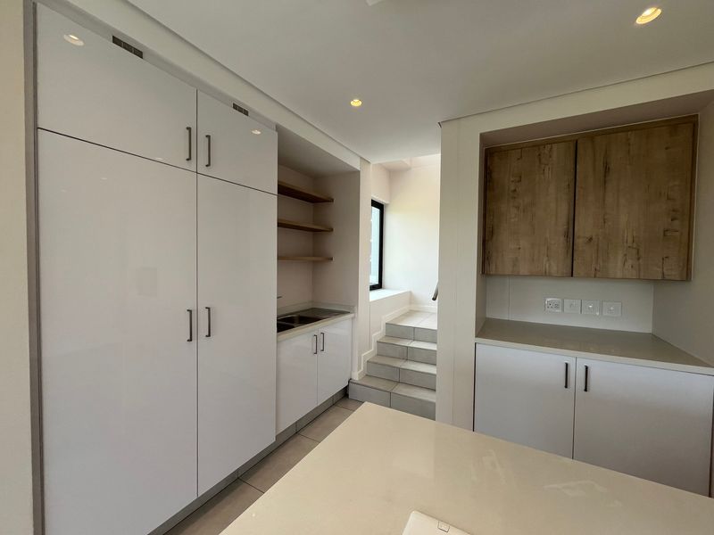 Modern scullery featuring sleek white and wood-grain cabinetry, stone countertops, a stainless steel sink, and recessed lighting, leading to a tiled staircase.