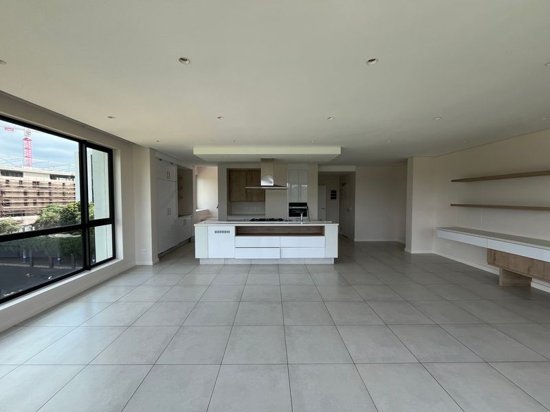 Spacious open-plan living and kitchen area featuring modern cabinetry, a central island with built-in appliances, tiled floors, and large windows with natural light.