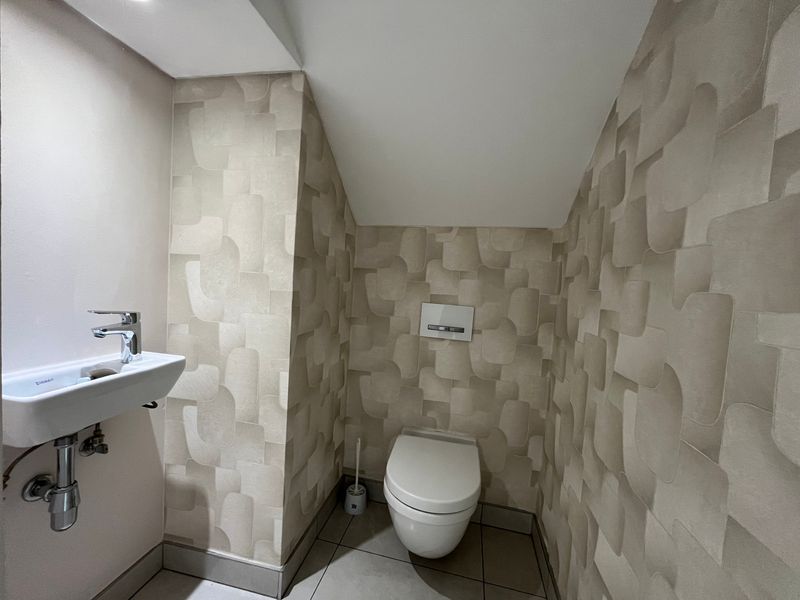 A compact, modern guest bathroom featuring a wall-mounted toilet with a concealed cistern, a sleek wall-mounted basin, and contemporary patterned wallpaper.