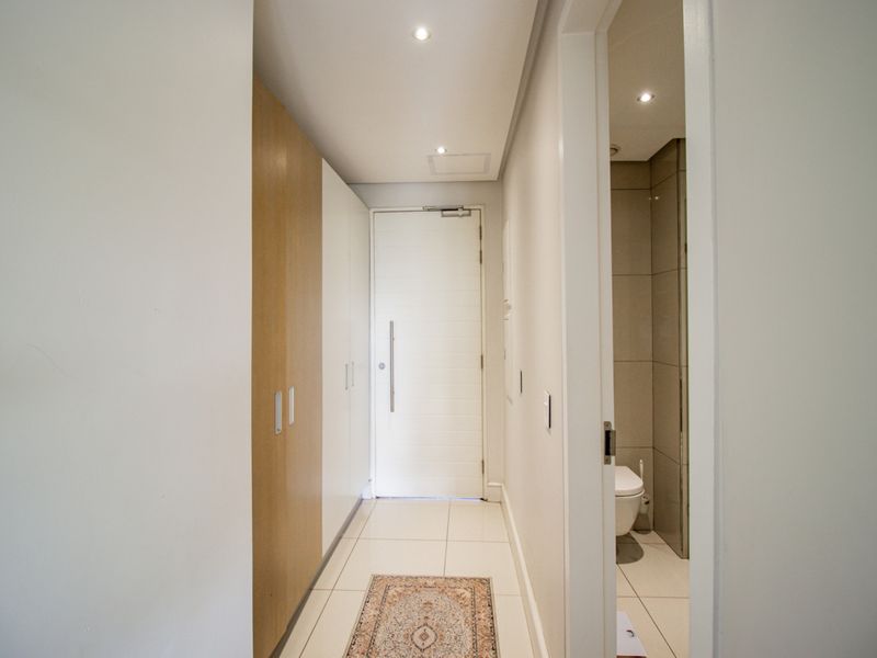 A clean and modern hallway featuring built-in storage cupboards, tiled flooring, and recessed downlighting, leading towards the main entrance and an adjacent bathroom.