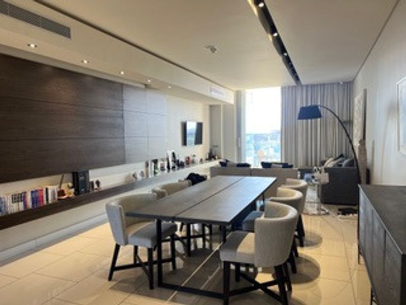 A modern open-plan living and dining area featuring sleek tiled floors, recessed ceiling lighting, and integrated wall paneling with built-in shelving.
