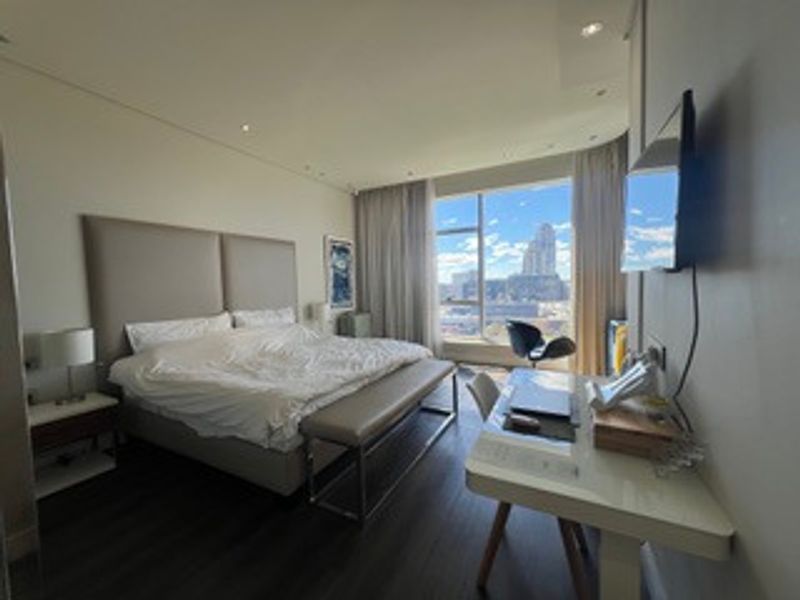 A modern bedroom featuring laminate flooring, recessed lighting, and a large window offering expansive city views.