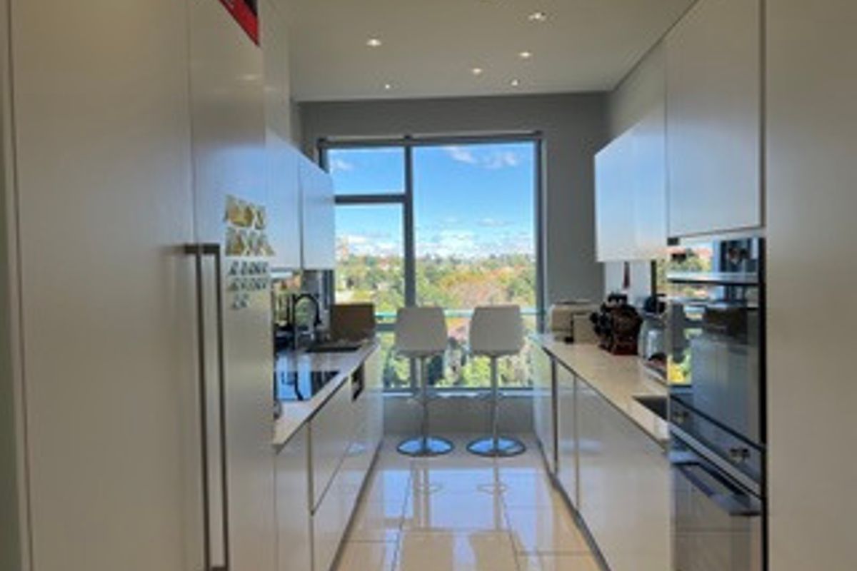 Modern galley-style kitchen featuring sleek white cabinetry, integrated appliances, tiled flooring, and a large window with natural light and scenic views.