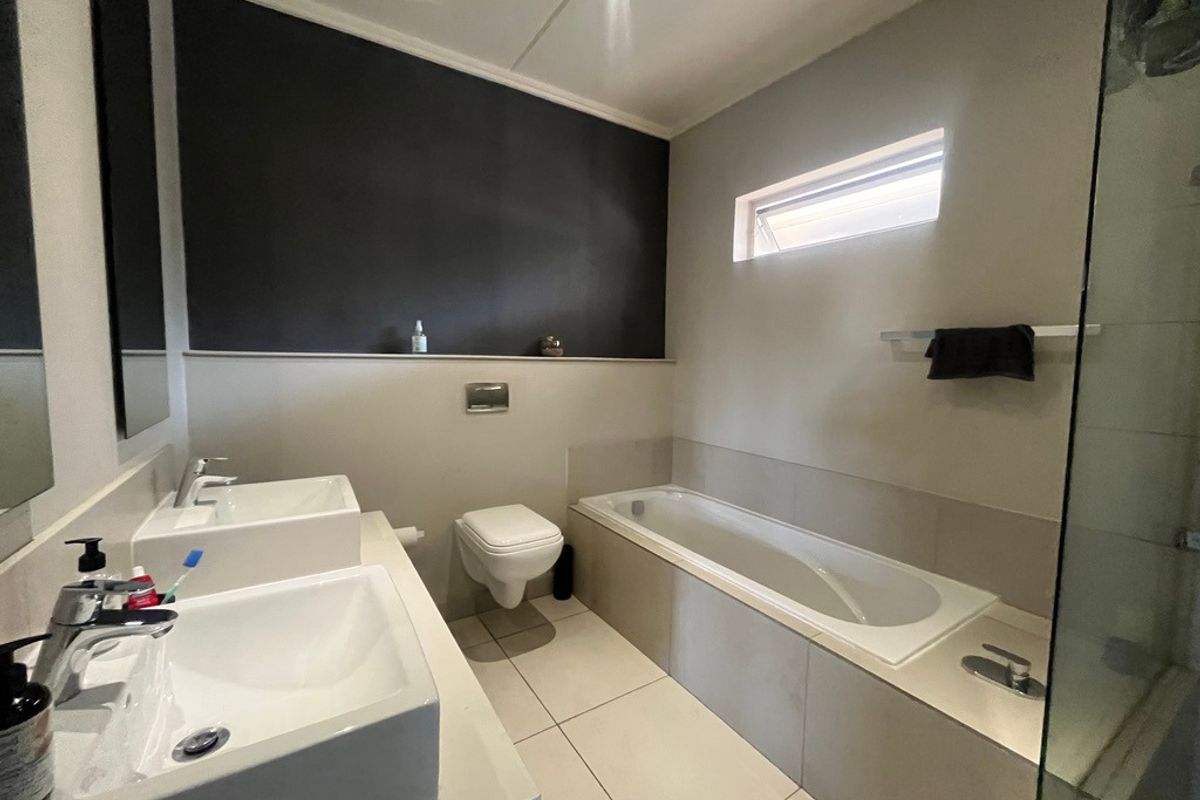Modern bathroom featuring a double vanity, wall-mounted toilet, built-in bathtub, and a separate glass-enclosed shower with neutral tiling.