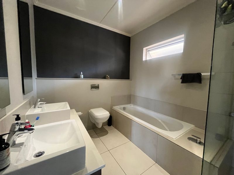 Modern bathroom featuring a double vanity, wall-mounted toilet, built-in bathtub, and a separate glass-enclosed shower with neutral tiling.