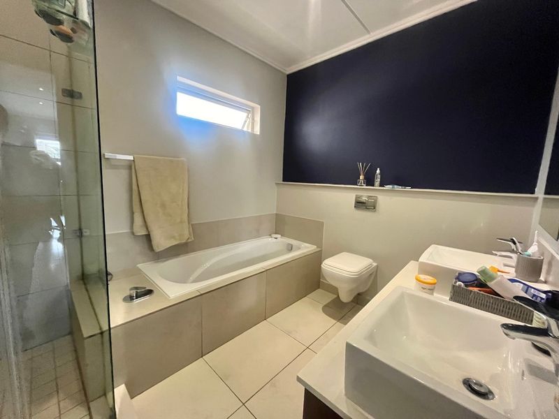 Modern bathroom featuring a double vanity, wall-mounted toilet, built-in bathtub, and a separate glass-enclosed shower with neutral tiling.