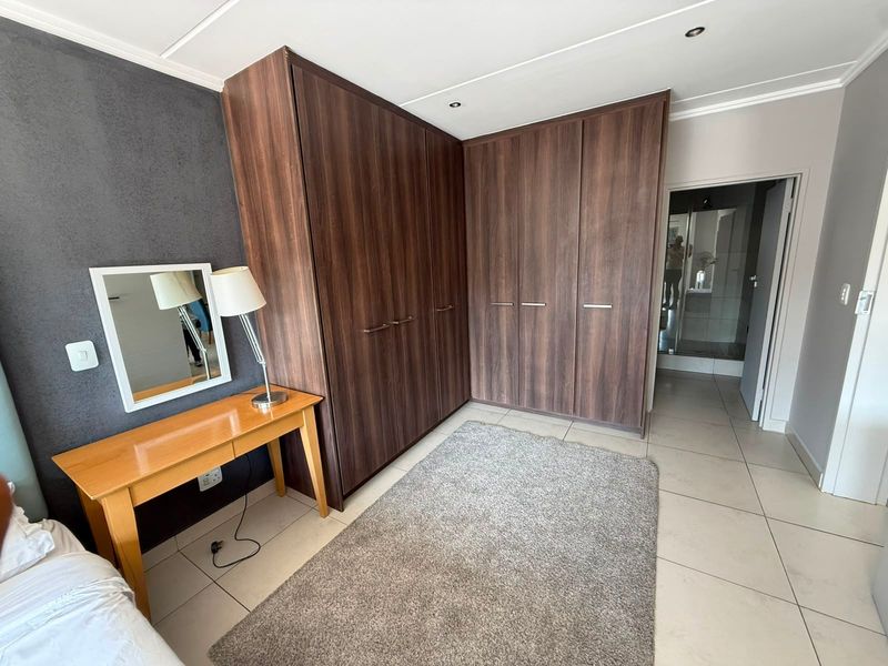 A well-maintained bedroom featuring dark wood built-in cupboards, neutral tiled flooring, and recessed lighting, with an adjoining en-suite bathroom.
