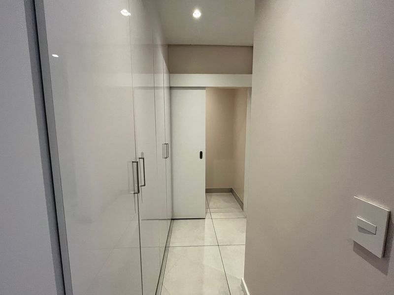 A clean, modern hallway featuring high-gloss built-in cupboards, neutral tiled flooring, and recessed downlighting, leading to a sliding door entrance.