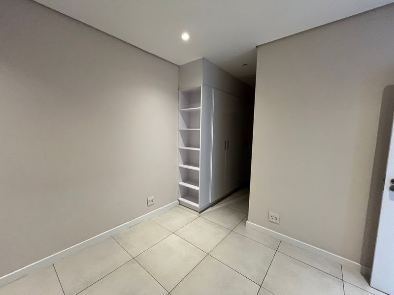 A clean, modern bedroom space featuring neutral walls, large format floor tiles, and integrated built-in cupboards with open shelving.