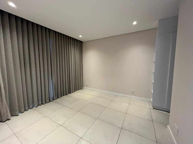 A bright, empty bedroom featuring neutral tiled flooring, recessed downlighting, and a built-in shelving unit, complemented by floor-to-ceiling curtains.