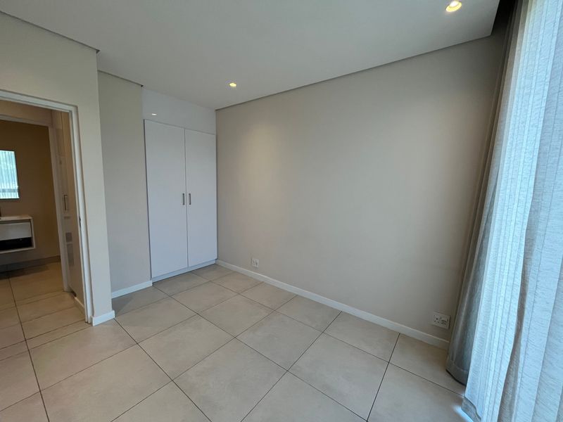 A clean, empty bedroom featuring neutral tiled flooring, recessed ceiling lighting, and a built-in wardrobe, with an adjacent view into a bathroom.