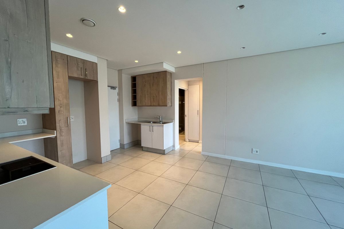 A modern, empty kitchen area featuring light wood-tone cabinetry, a built-in hob, a sink, and neutral tiled flooring with recessed ceiling lighting.