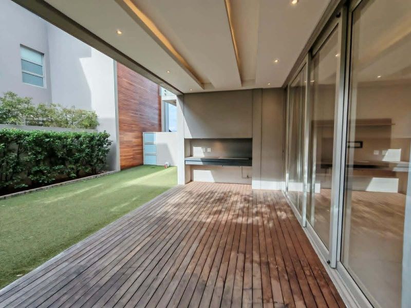 A modern covered patio featuring timber decking, a built-in braai, and seamless access to the interior through large sliding glass doors.