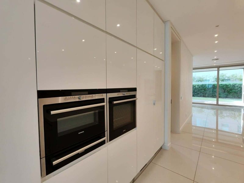 Modern kitchen area featuring high-gloss white cabinetry, integrated Siemens ovens, and sleek tiled flooring, leading to an open-plan living space with garden access.