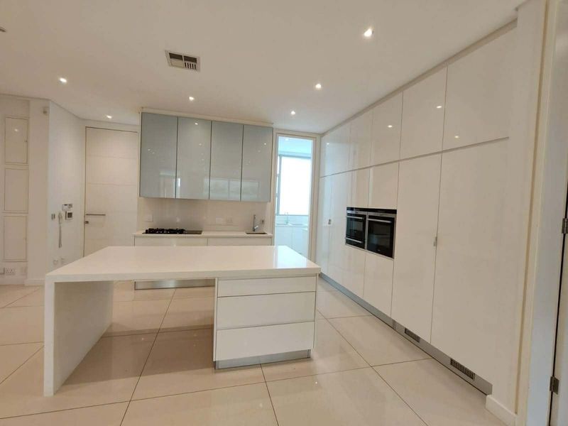 Modern, minimalist kitchen featuring high-gloss white cabinetry, a central island with stone countertops, integrated appliances, and sleek tiled flooring.