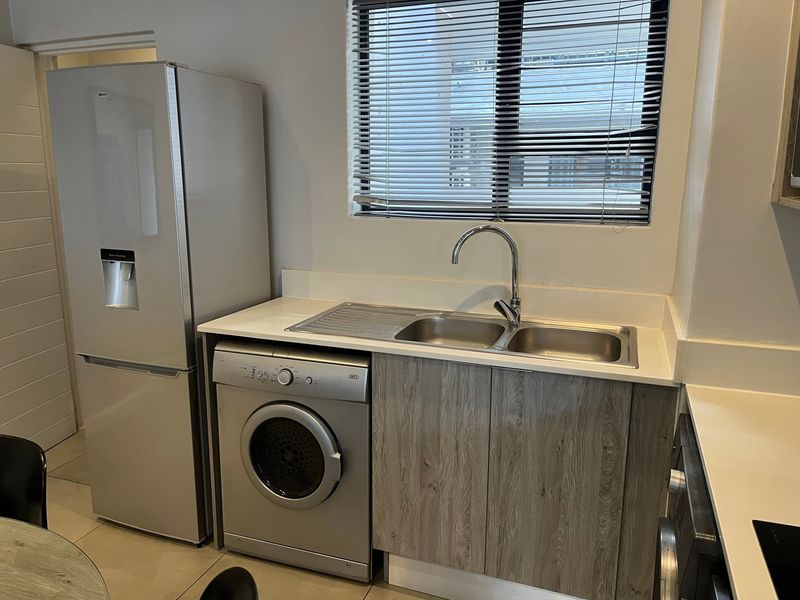 A functional laundry area featuring a double stainless steel sink, stone countertop, and modern laminate cabinetry with space for a washing machine.