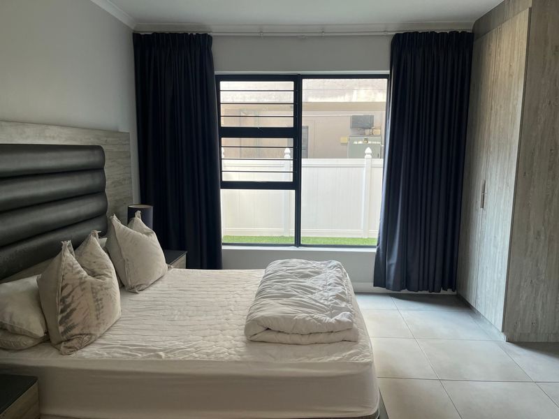 A well-maintained bedroom featuring neutral tiled flooring, built-in cupboards, and a window with security bars overlooking an outdoor area.