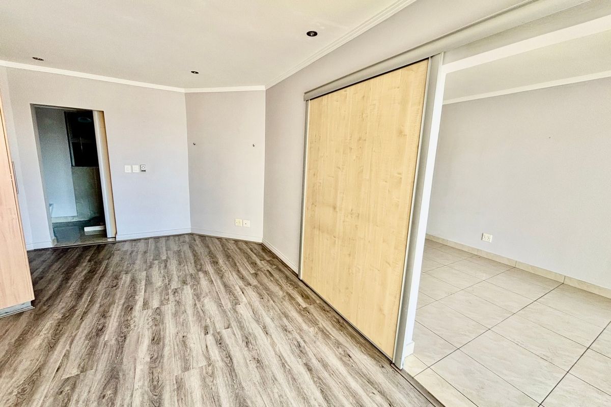 A bright, empty bedroom featuring a built-in sliding door wardrobe, a mix of laminate and tiled flooring, and modern recessed downlighting.