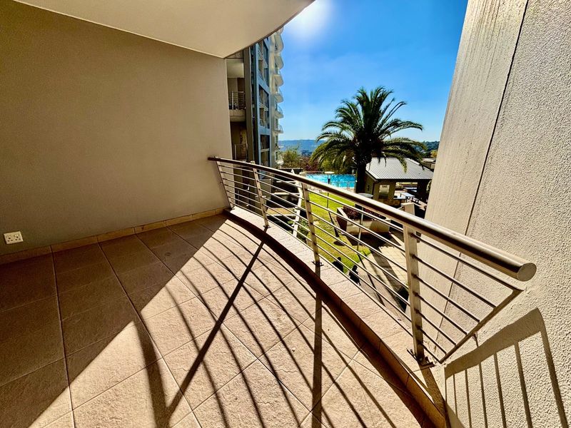A spacious covered balcony featuring tiled flooring and a modern stainless steel balustrade, offering views of the complex pool and garden area.
