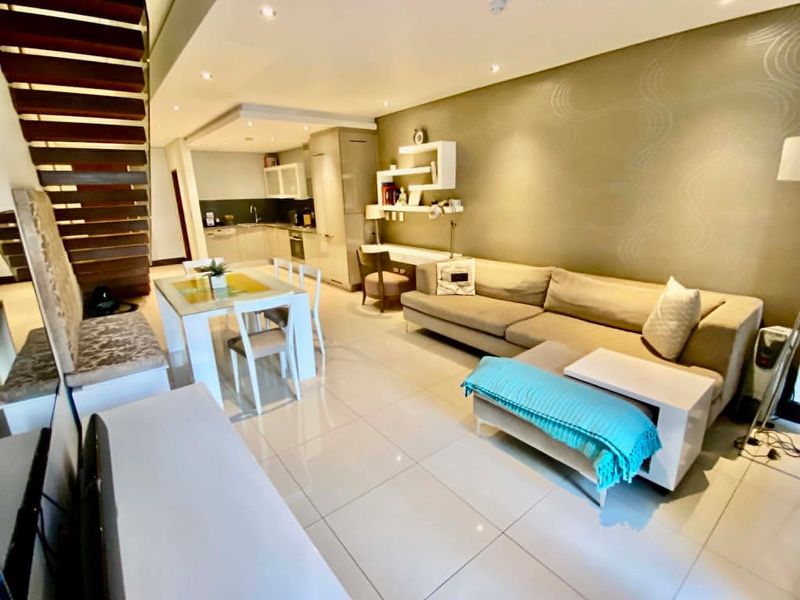 Spacious open-plan living and dining area featuring a modern kitchen, built-in desk, and a striking open-tread staircase with polished tiled flooring throughout.