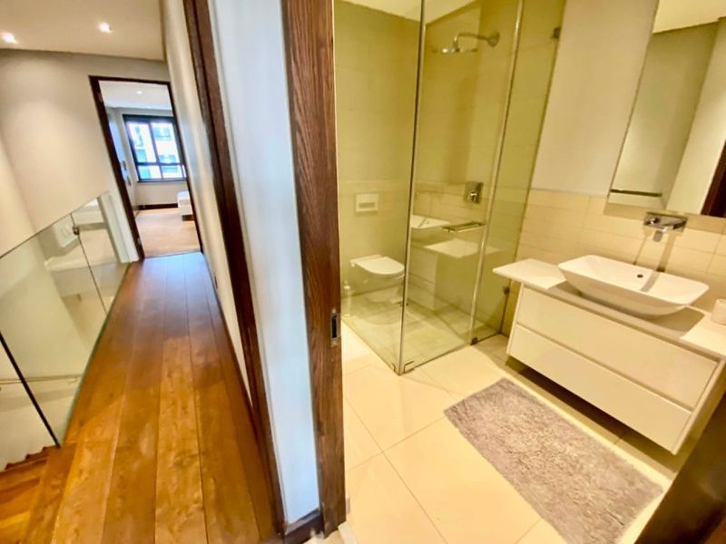 Modern bathroom featuring a vessel sink vanity, glass-enclosed shower, and wall-mounted toilet, adjacent to a wooden-floored hallway.