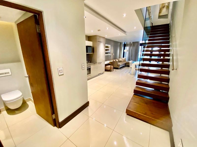 Modern open-plan apartment featuring a sleek kitchen, tiled flooring, and a striking wooden staircase with a glass balustrade leading to the upper level.