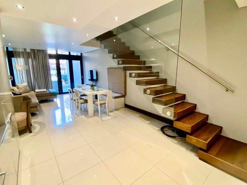 Modern open-plan living area featuring a striking floating timber staircase with glass balustrade, polished floor tiles, and large sliding doors leading to the exterior.