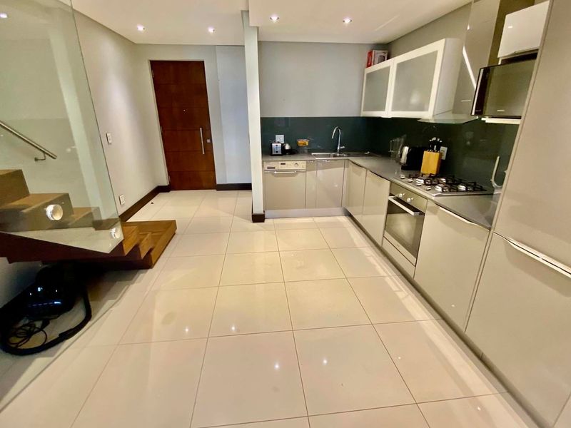 Modern kitchen featuring high-gloss cabinetry, integrated appliances, gas hob, and sleek glass splashbacks, seamlessly connected to an open-plan living area with timber staircase.