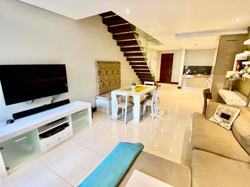 Modern open-plan living and dining area featuring polished floor tiles, a striking open-tread staircase with glass balustrade, and an integrated kitchen.