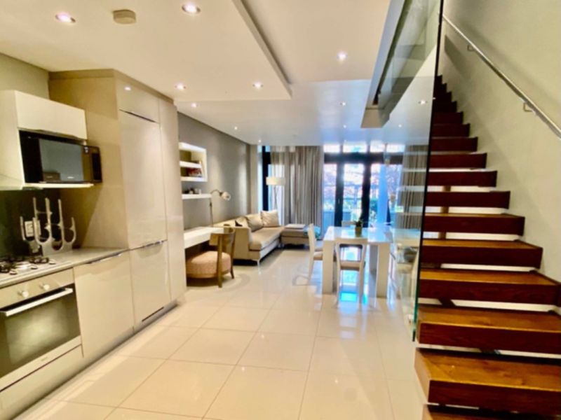 Modern open-plan living area featuring a sleek kitchen with integrated appliances, tiled flooring, and a striking timber staircase with glass balustrade.