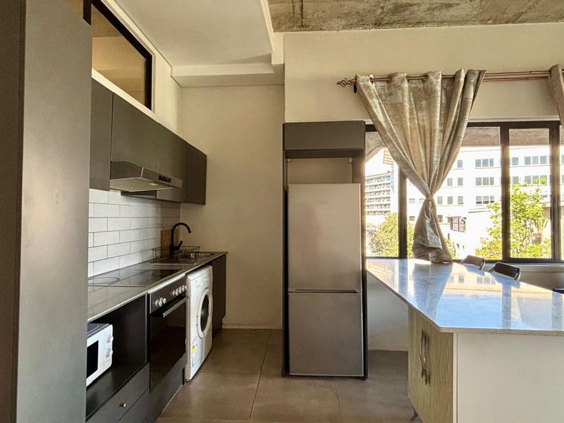 Modern open-plan kitchen featuring dark cabinetry, stone countertops, subway tile backsplash, and an exposed concrete ceiling, seamlessly integrated with the living area.