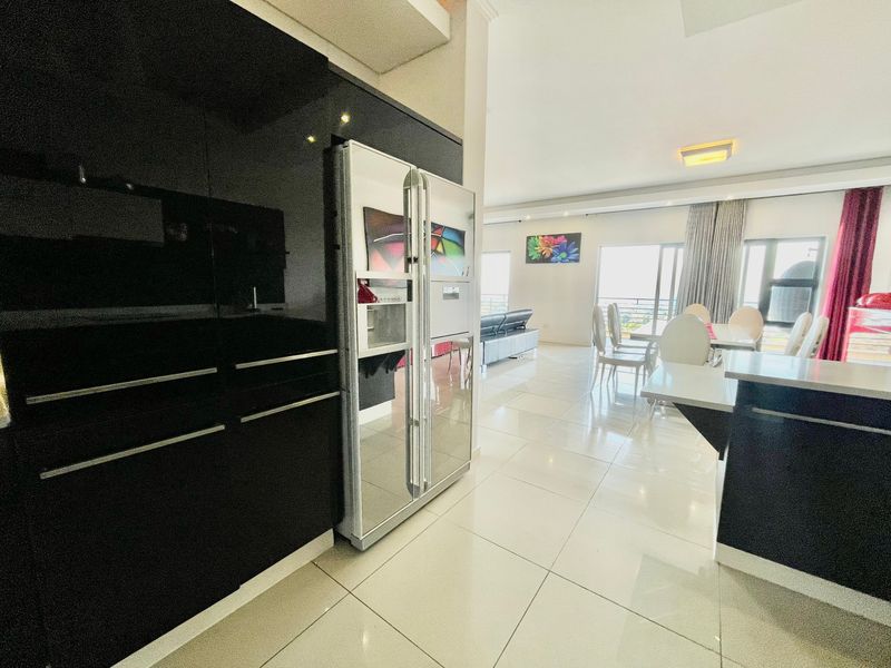 Modern open-plan living area featuring high-gloss black kitchen cabinetry, integrated appliances, and bright tiled flooring leading to a balcony with natural light.