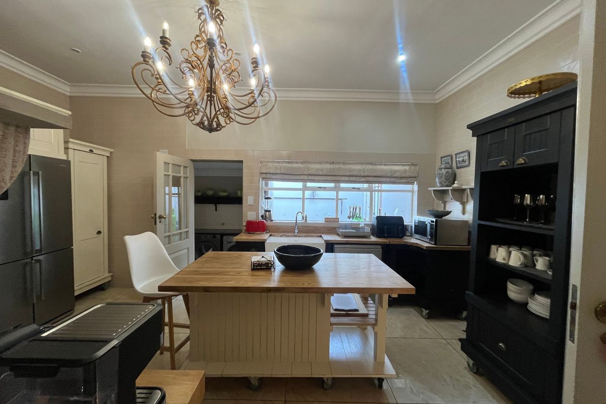 A charming kitchen featuring a central wooden island, farmhouse sink, tiled backsplash, and elegant chandelier lighting, with an adjacent scullery area.