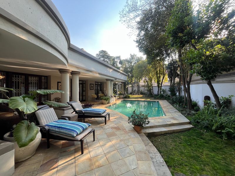 A well-maintained outdoor pool area featuring a stone-paved patio, covered veranda with architectural columns, and a landscaped garden setting.