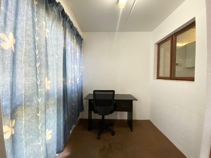 A small office space featuring carpet flooring and an internal window looking into the adjacent room.
