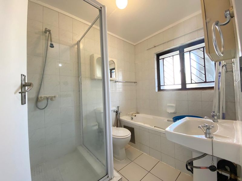 A functional bathroom featuring a separate shower enclosure, bathtub, pedestal basin, and toilet, with tiled finishes and security bars on the window.