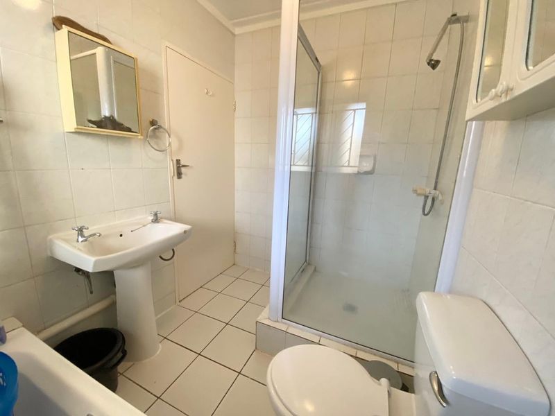 A functional bathroom featuring a pedestal basin, a glass-enclosed shower, and a standard toilet with white wall and floor tiling.