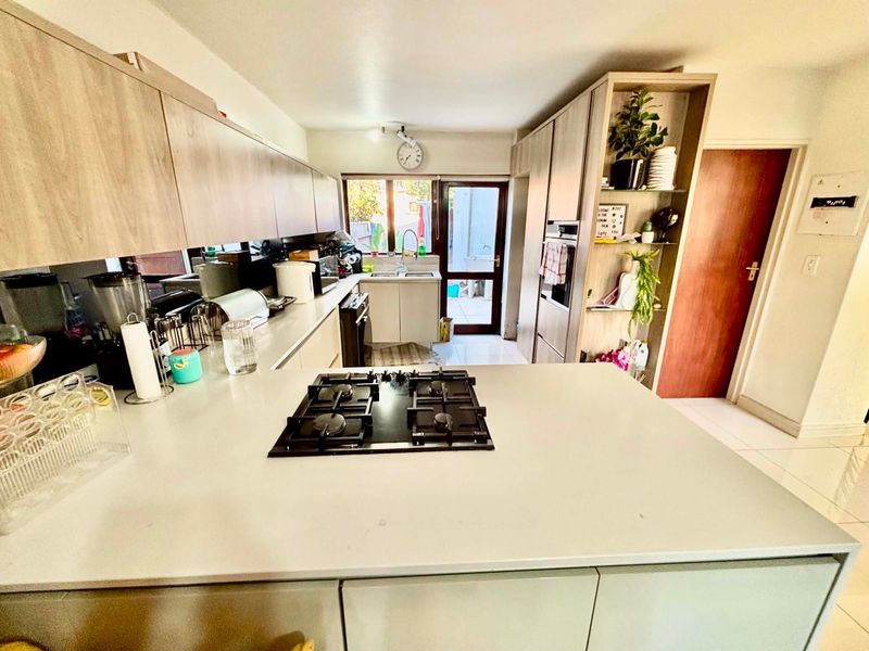 Modern kitchen featuring light wood cabinetry, stone countertops, a central gas hob, built-in oven, and a mirrored splashback with direct access to the rear exterior.