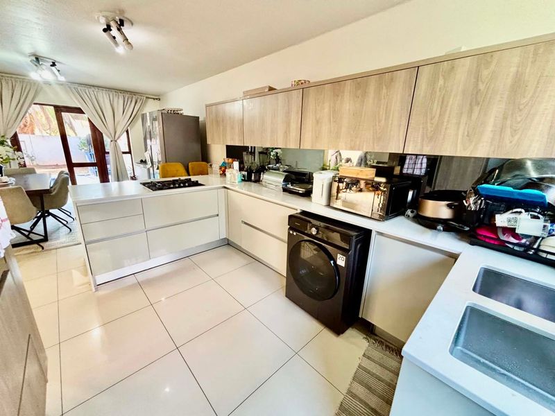 Modern kitchen featuring light wood-grain upper cabinetry, sleek white base units, stone countertops, a gas hob, and a mirrored splashback with tiled flooring.