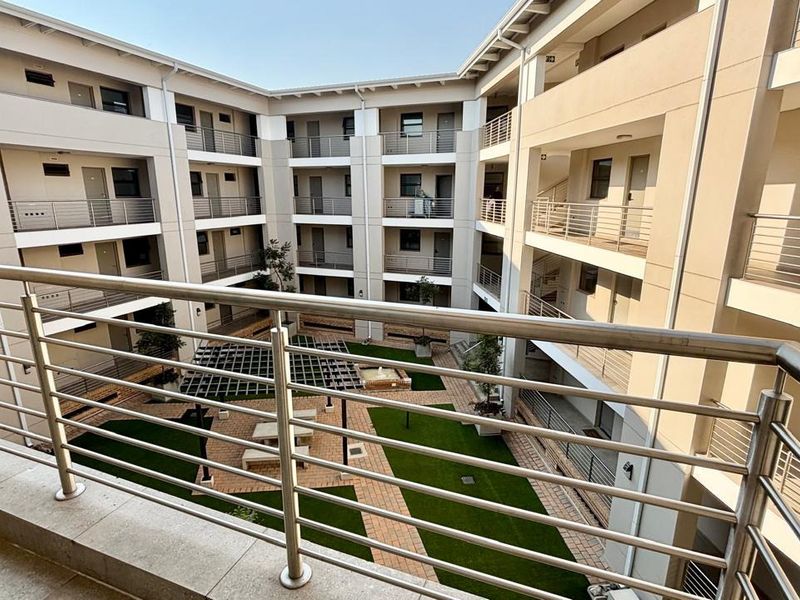 View from a balcony overlooking a well-maintained apartment complex courtyard featuring paved walkways, artificial lawn, and a pergola.