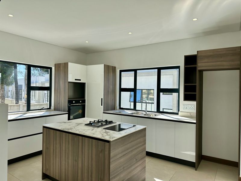 Modern kitchen featuring a central island with gas cooktop, built-in oven, stone countertops, and ample cabinetry, illuminated by recessed lighting and large windows.