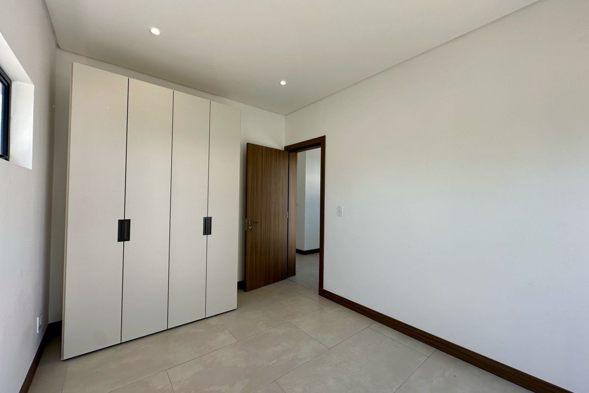 A bright, modern bedroom featuring neutral tiled flooring, recessed downlighting, and a large built-in wardrobe with clean, contemporary finishes.