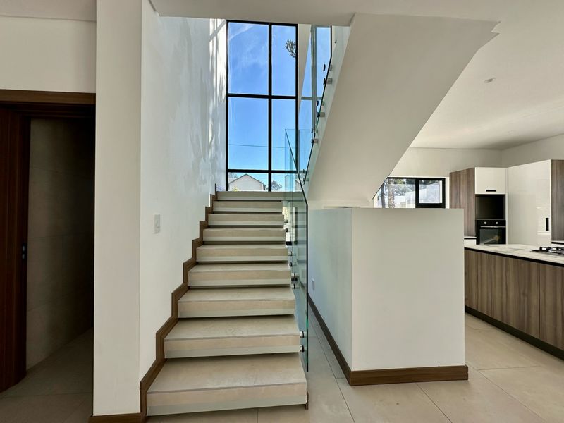 Modern open-plan interior featuring a striking staircase with glass balustrade, large floor-to-ceiling window, and adjacent contemporary kitchen area.