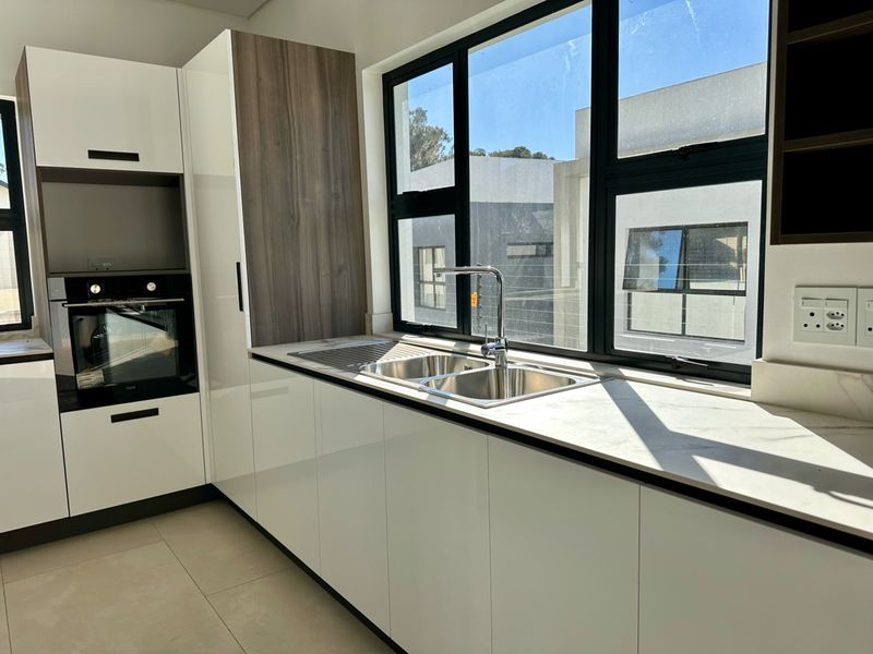 Modern kitchen featuring high-gloss cabinetry, stone countertops, a stainless steel double sink, and a built-in oven with large windows for natural light.